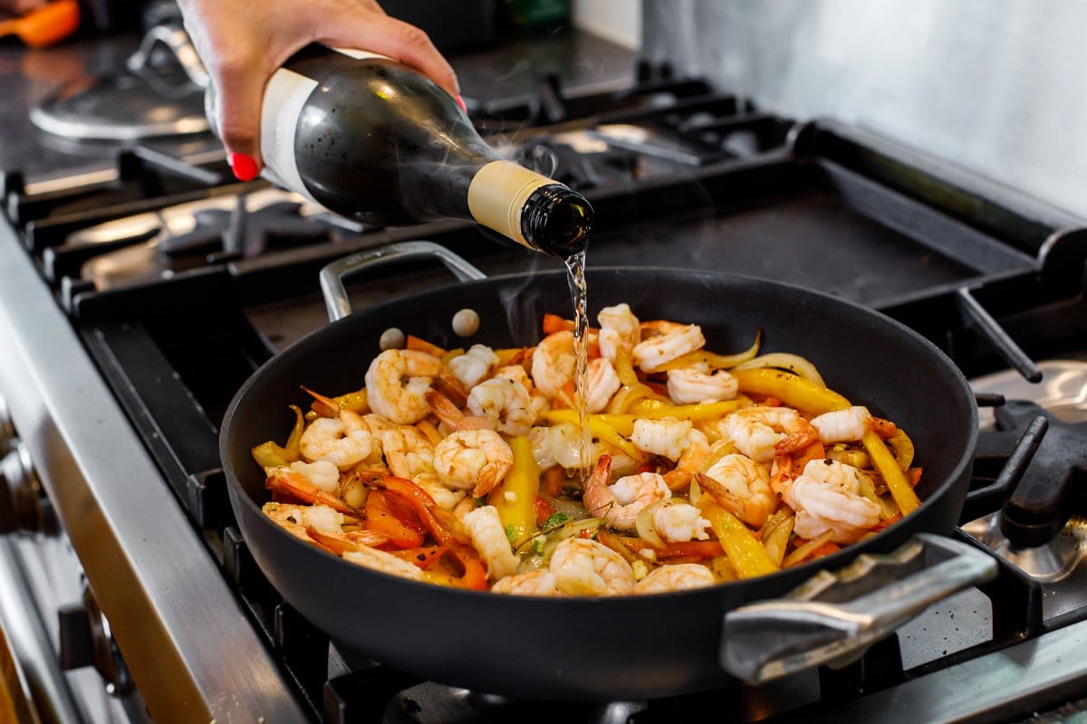 Pouring white wine into skillet with sautéed shrimp and peppers.