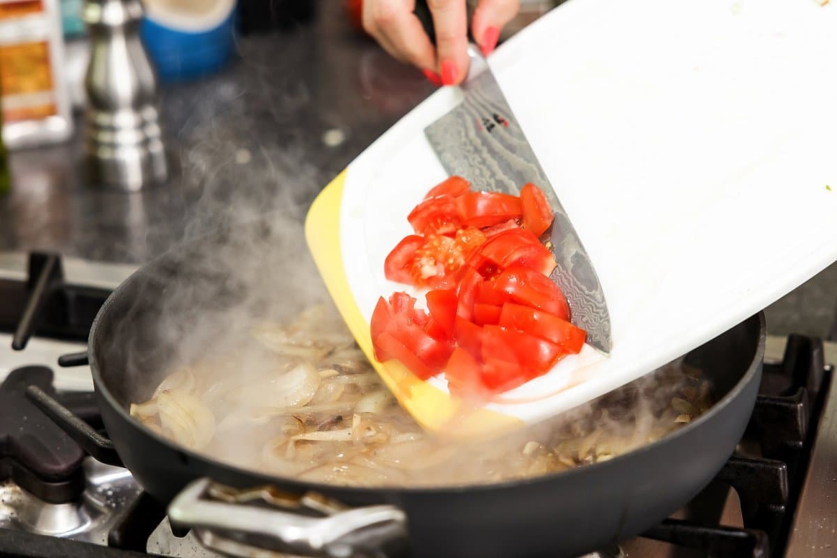 Tomatoes being added to a skillet with cooked onion.