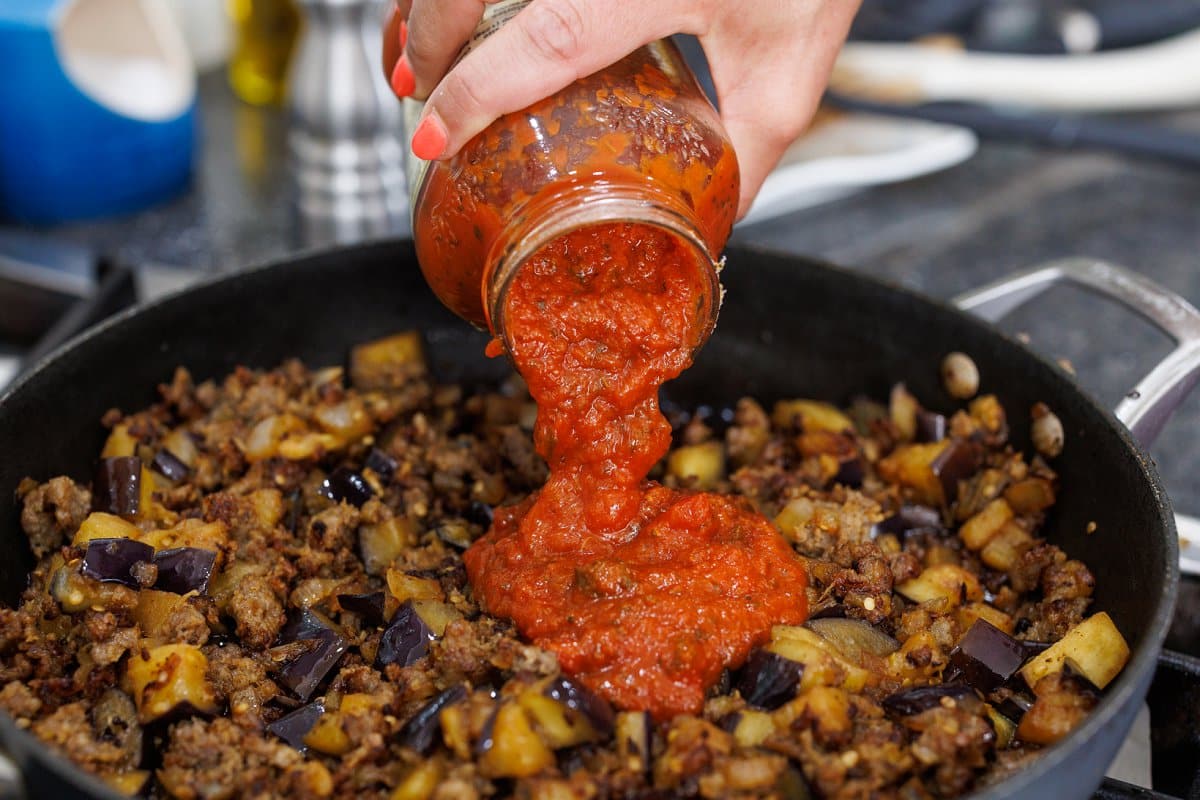 Pouring jarred tomato sauce into skillet with eggplant and sausage.