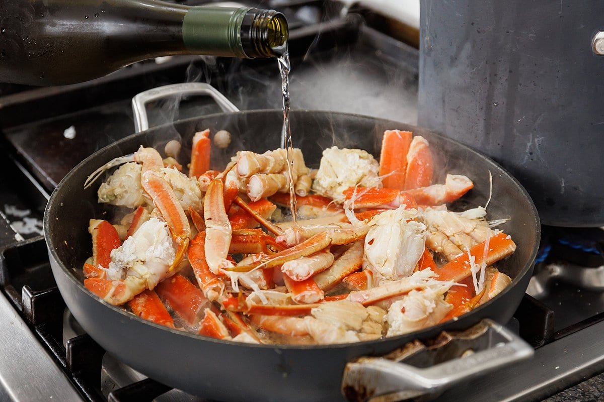 Pouring white wine over crab legs in a large skillet.
