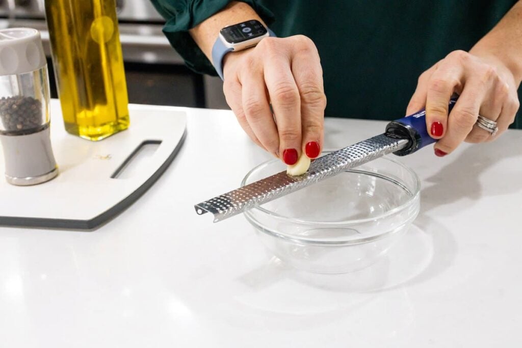 Grating a garlic clove into a small glass bowl with a microplane.