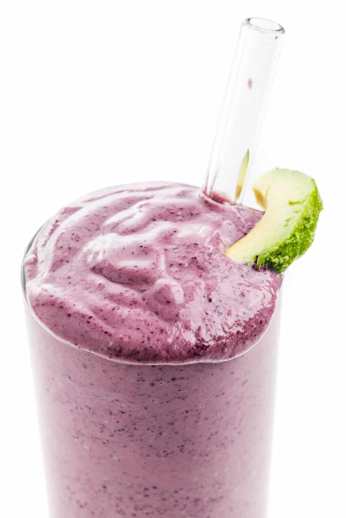 Close up of the top of an avocado blueberry smoothie with a slice of avocado and a clear straw.