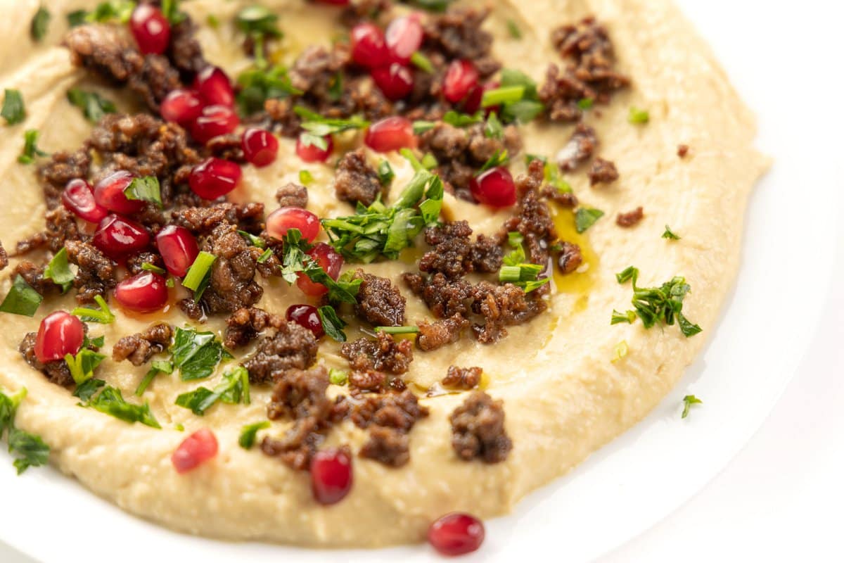 Warm hummus with ground lamb and pomegranate on a large white platter.