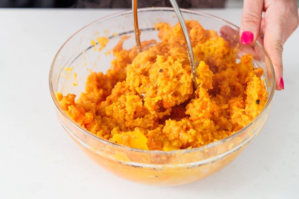 Using a potato masher to mash the cooked butternut squash and sweet potatoes in a glass bowl.