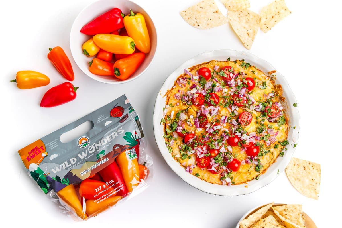 Baked hummus dip in a white bowl next to a bowl of wild wonders mini peppers and their packaging.