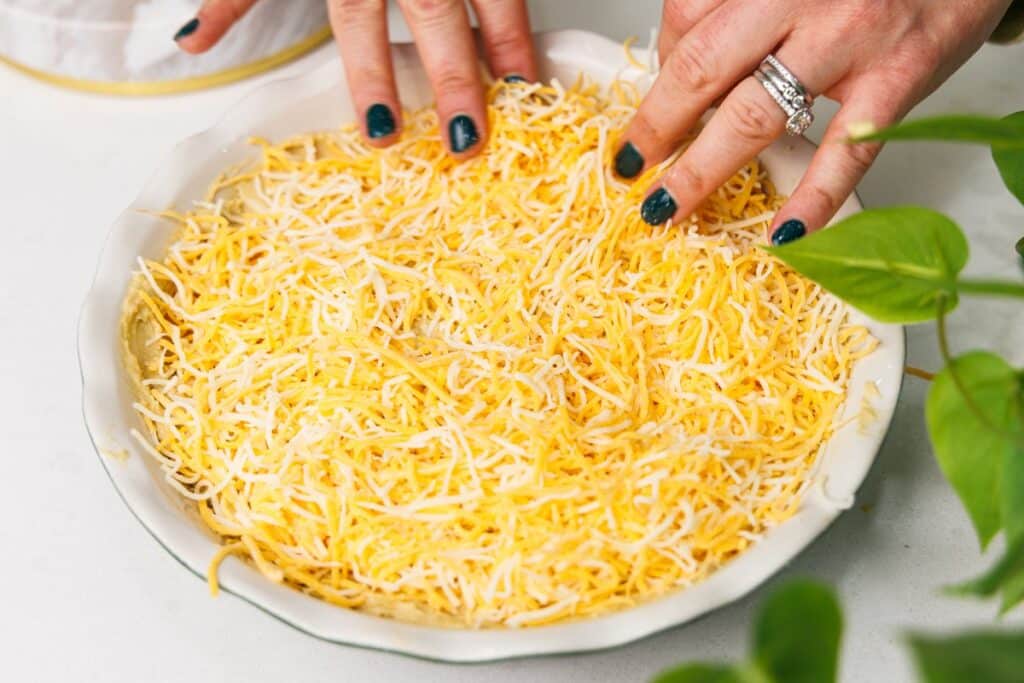 Spreading shredded cheese over the top of hummus in a baking dish.