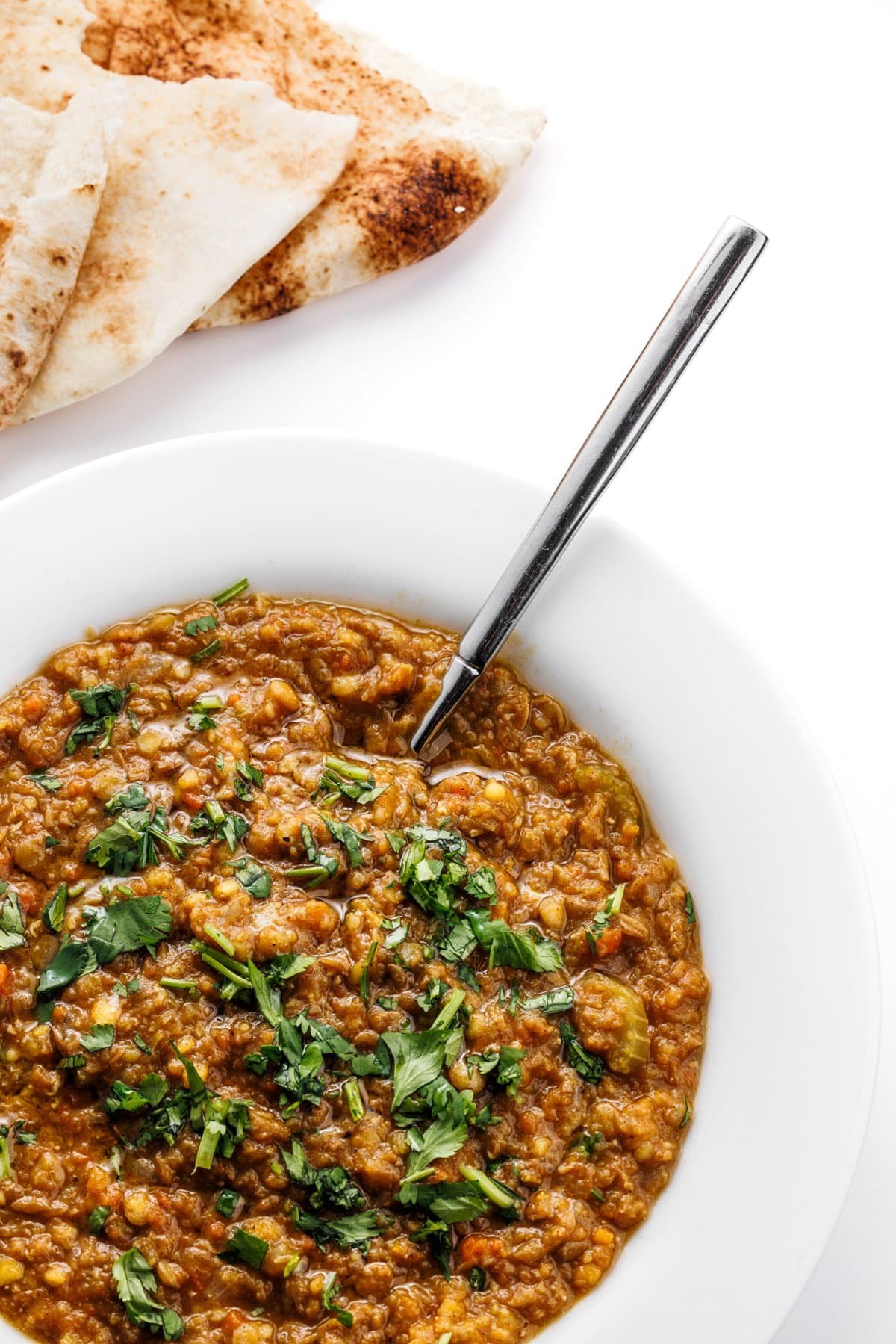 Spoon sitting in a bowl of Arabic lentil soup in a white bowl next to pita bread.