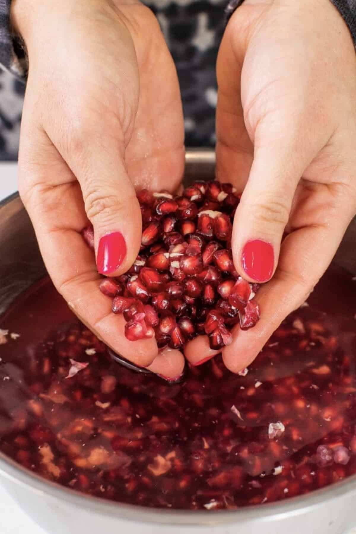 Two hands holding a bunch of pomegranate seeds over a metal bowl with more seeds.