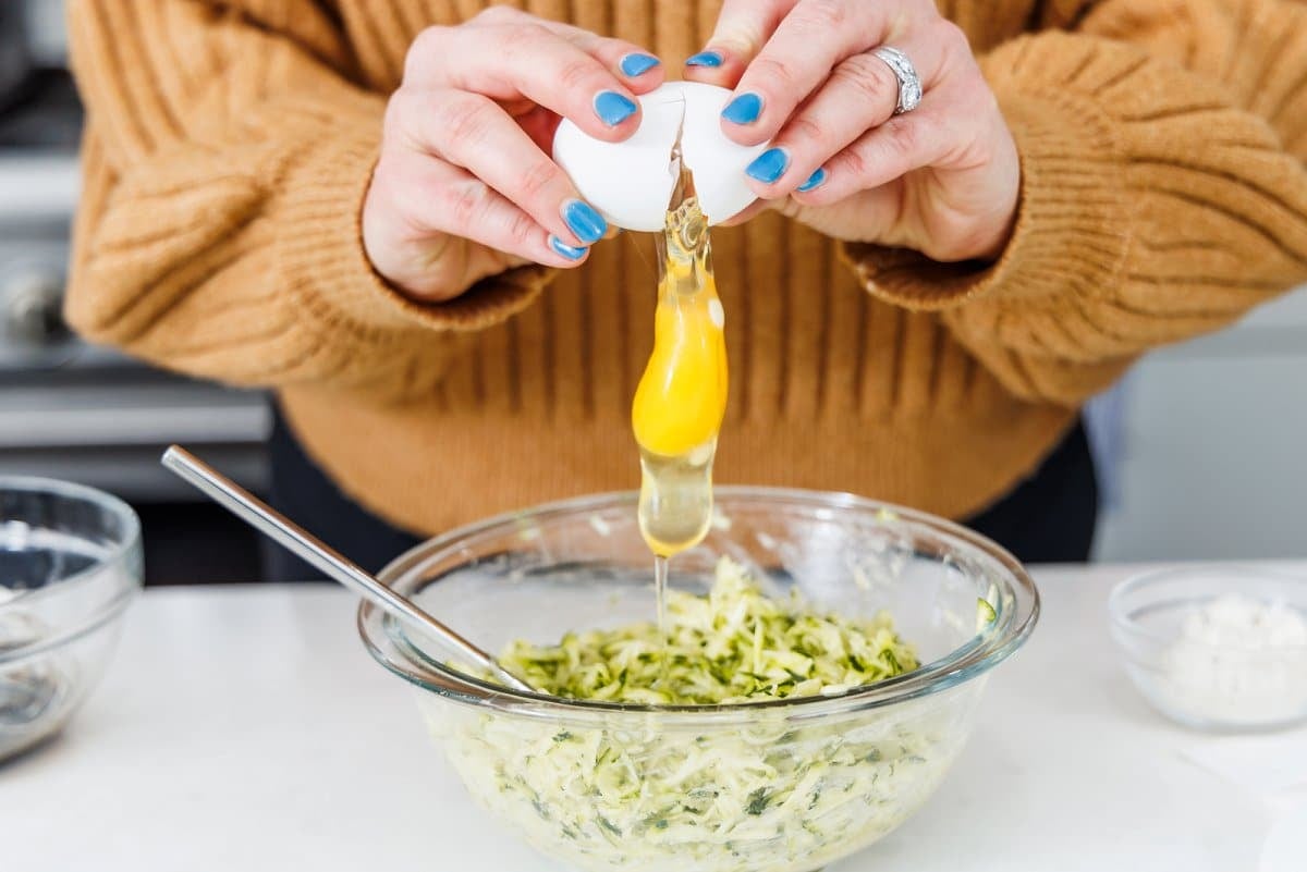 Cracking an egg into a glass bowl filed with shredded zucchini.