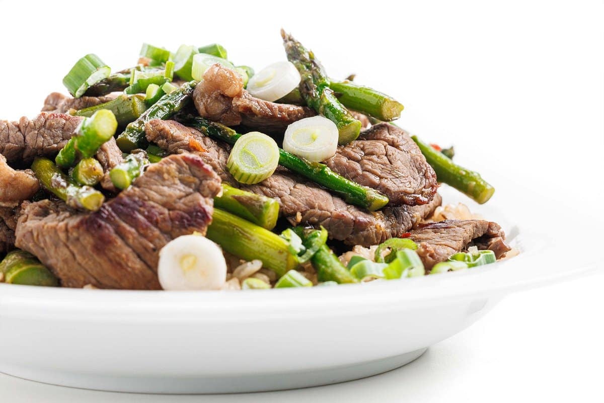 Edge of a white plate topped with rice and ginger beef and asparagus stir fry.