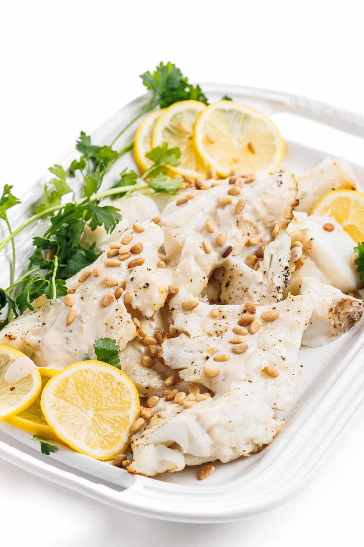Grilled white fish with tahini lemon sauce on a white platter with pine nuts, lemon slices, and parsley.