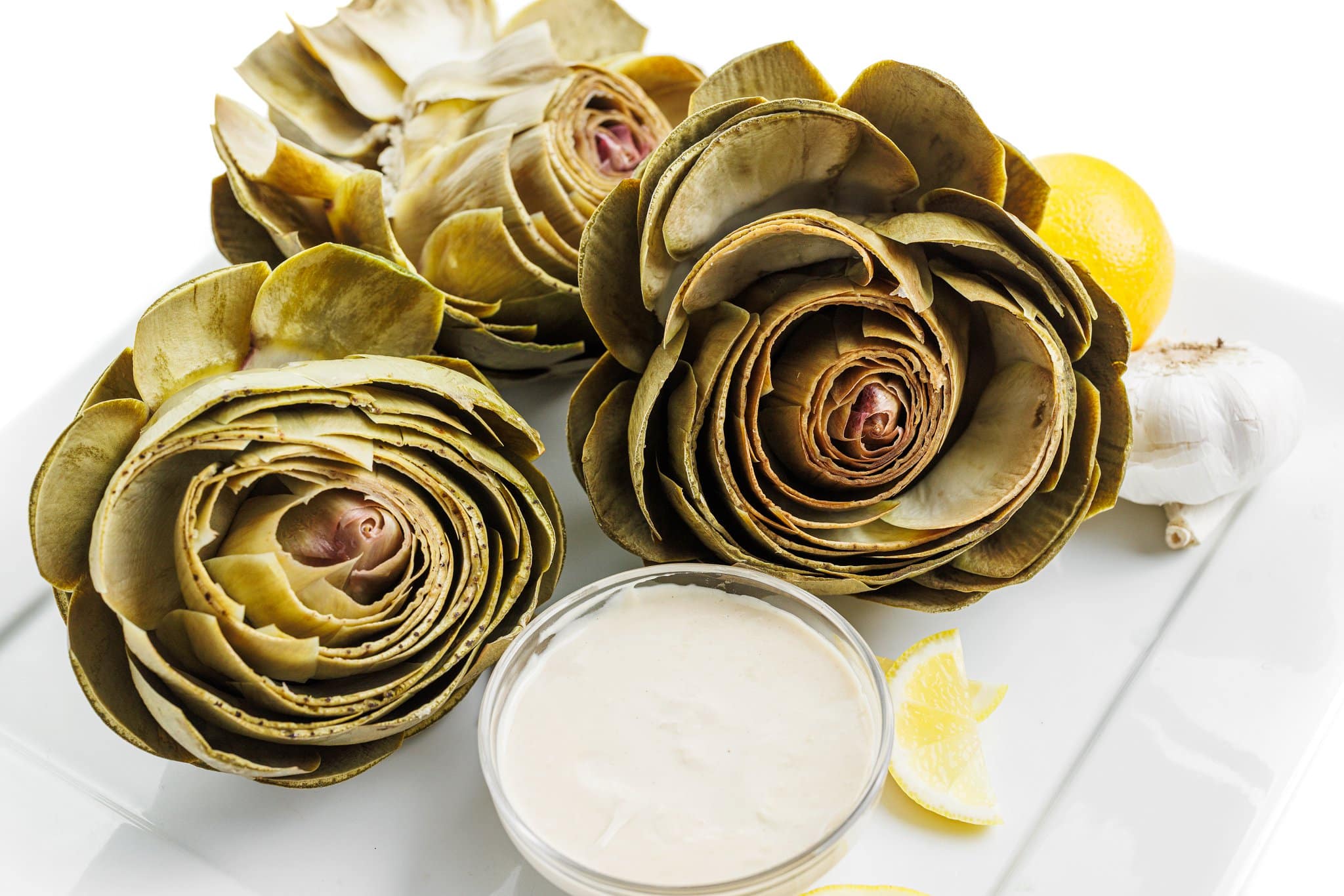 Three artichokes on a white plate with tahini dipping sauce.