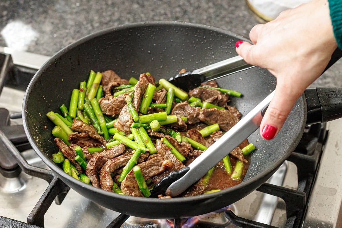 Tossing beef and asparagus in stir-fry sauce in wok on stove.