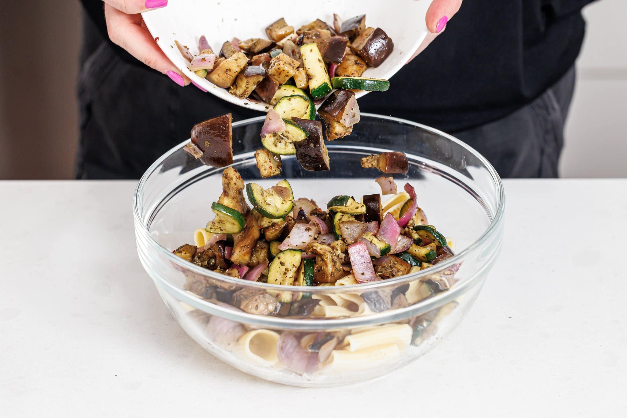 Adding grilled veggies to a bowl with cooked pasta.