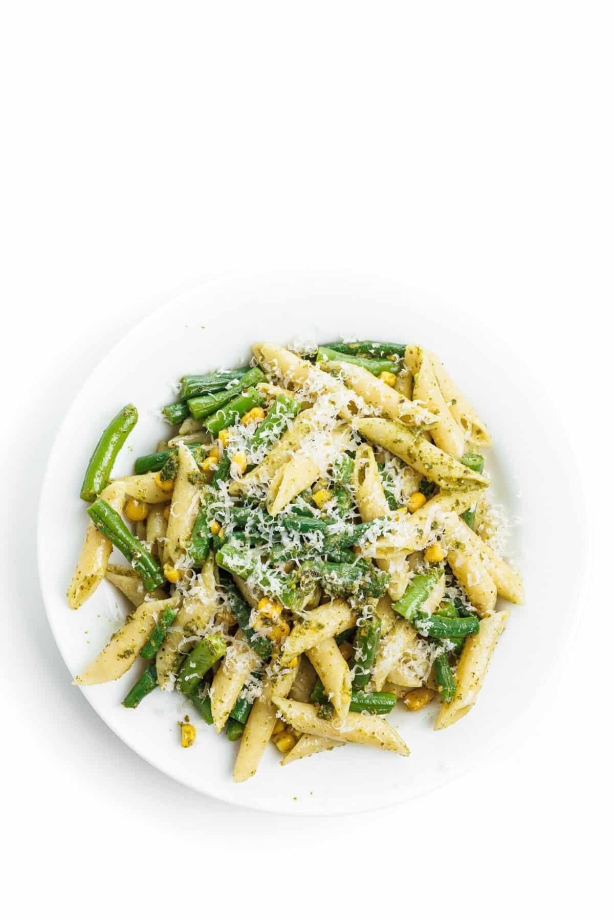 A round white plate with pesto pasta salad with green beans and corn on it.