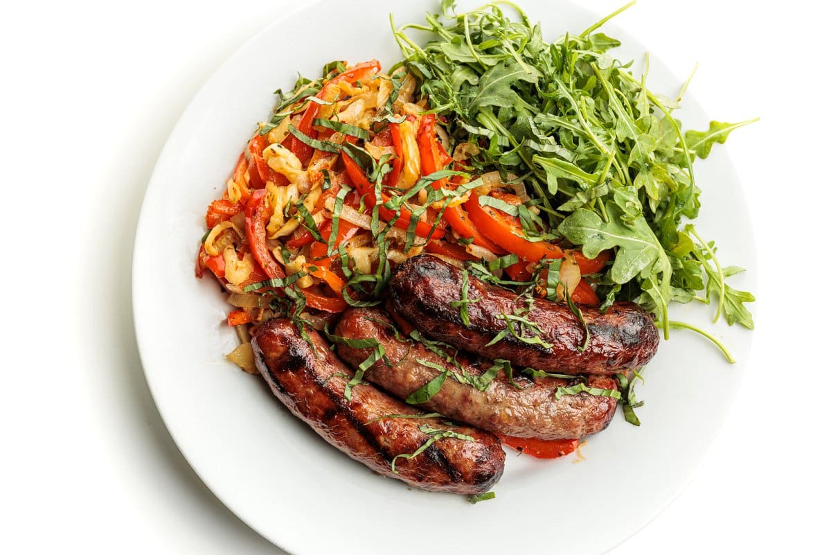 Grilled bratwurst with peppers and onions on a plate with arugula.