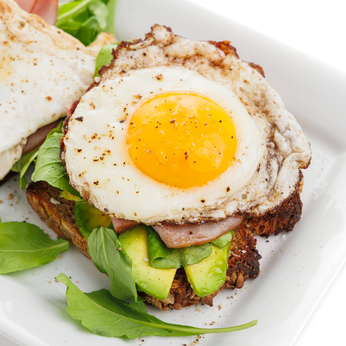 Avocado toast with egg, ham, and arugula on a white plate.