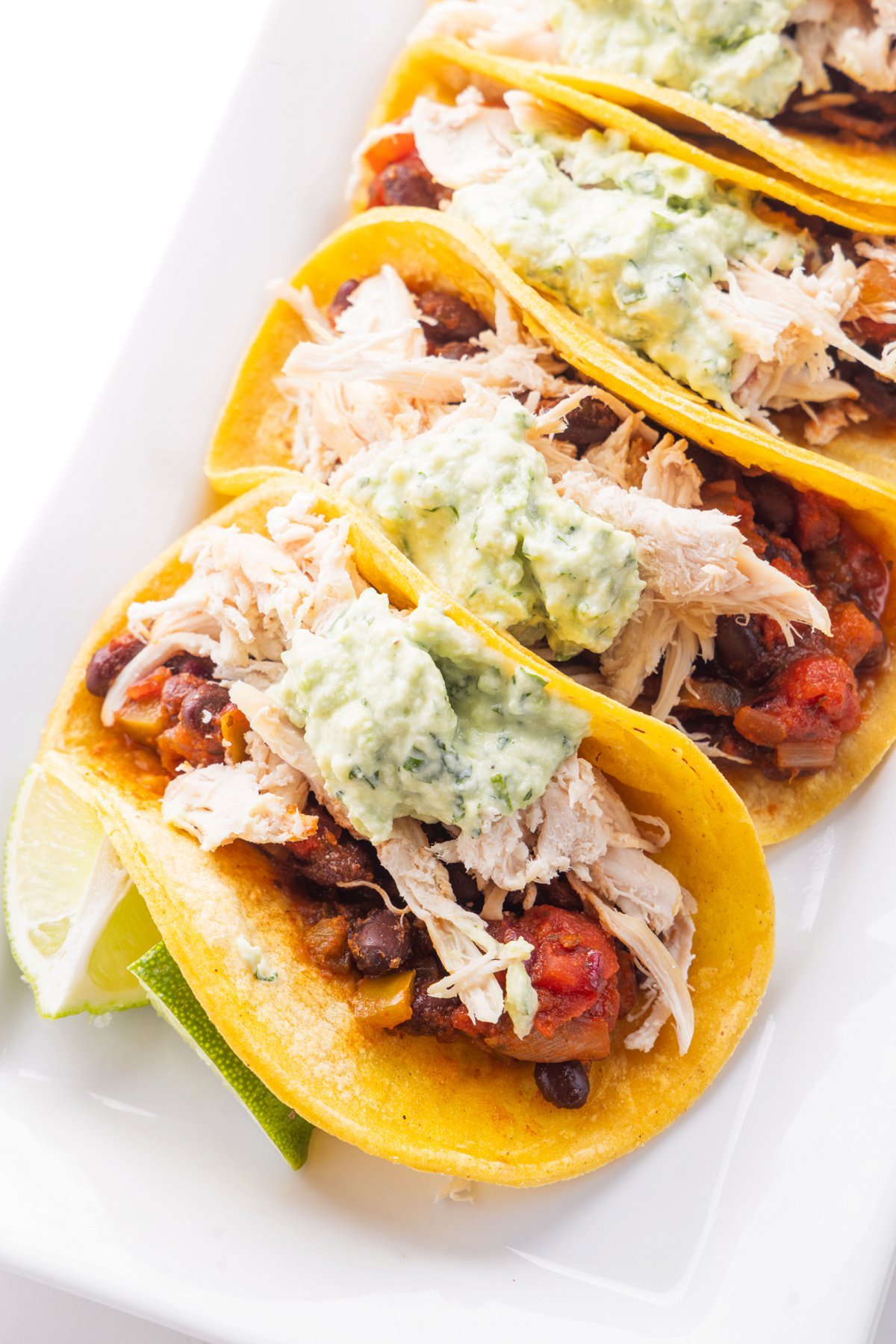 Four chicken and black bean tacos topped with avocado crema on a white plate.